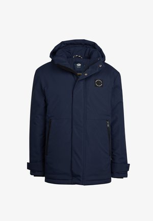 Chaqueta aislante de color navy con capucha, cremallera frontal y dos bolsillos laterales. Lleva puesta una camiseta de cuello alto verde y está combinada con vaqueros azules y zapatillas blancas.