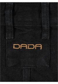 Melna dūnu audums ar uzšuvumu, kurā izšūts zelta krāsā zīmola nosaukums "DADA". Redzamas dūriens un jostas detaļas.