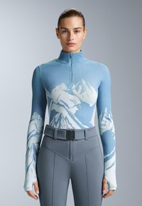 Bluza dolgih rokavov z zadrgo v svetlo modri barvi, ki prikazuje gorski vzorec v beli barvi, v kombinaciji z oprijetimi sivo visokimi hlačami in pasom.