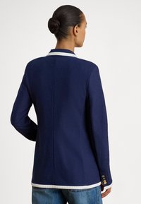 Mulher de cabelo escuro apanhado num coque usa um blazer azul-marinho com remate branco, botões dourados nos punhos e jeans azuis, vista de costas.
