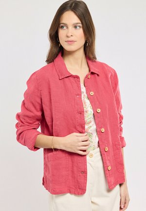 Femme aux cheveux bruns portant une veste rouge à manches longues boutonnée sur un haut à motifs et un pantalon de couleur claire, regardant sur le côté.