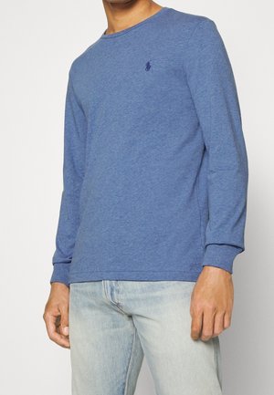 Mann trägt ein langärmliges blaues Rundhals-T-Shirt mit kleinem gesticktem Logo auf der Brust und hellblaue Jeans, steht vor weißem Hintergrund.