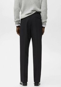 Uomo che indossa pantaloni neri su misura, maglione grigio lavorato a maglia, polsini bianchi della camicia visibili e scarpe eleganti nere, in piedi davanti a uno sfondo semplice.