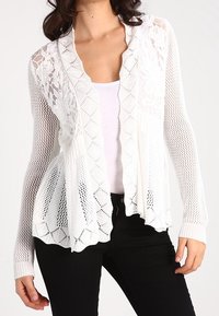 Vit, öppenstickad cardigan med ett blomstermönster i spets, långa ärmar och en lös passform. Buren över en ljus topp och svarta byxor.