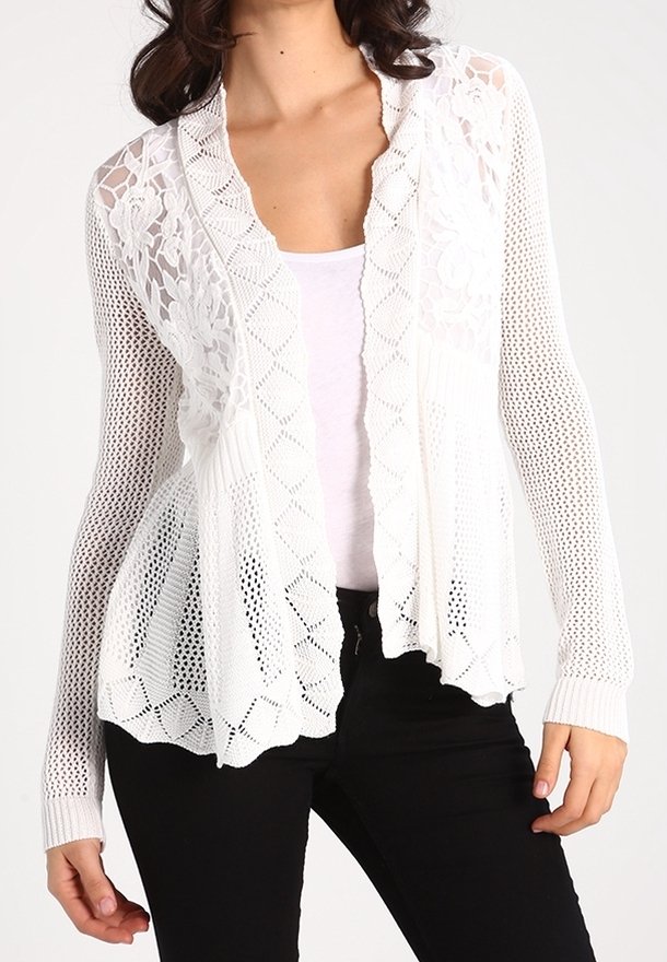 Vit, öppenstickad cardigan med ett blomstermönster i spets, långa ärmar och en lös passform. Buren över en ljus topp och svarta byxor.