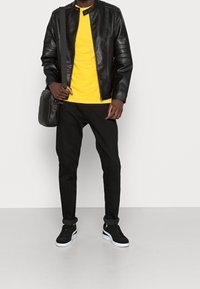 Giacca di pelle nera con spalle a coste, t-shirt gialla, jeans neri e sneakers nere con suola bianca, che tiene in mano una borsa a tracolla nera.