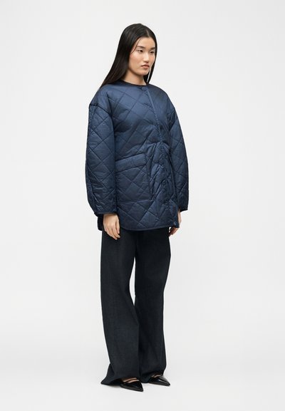 Γυναίκα με μπλε ναυτικό καπιτονέ μπουφάν με μπροστινές τσέπες, μαύρο παντελόνι καμπάνα και μαύρα μυτερά παπούτσια, στέκεται μπροστά σε ένα απλό φόντο.