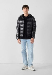 Giacca a vento nera con finitura lucida, dotata di zip frontale, maniche aderenti e cappuccio, abbinata a jeans azzurri e sneakers grigie.