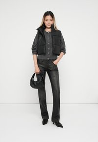 Gilet imbottito nero sopra un cardigan grigio, jeans larghi grigi e tacchi neri. Presenta anche una borsa nera, su uno sfondo neutro.