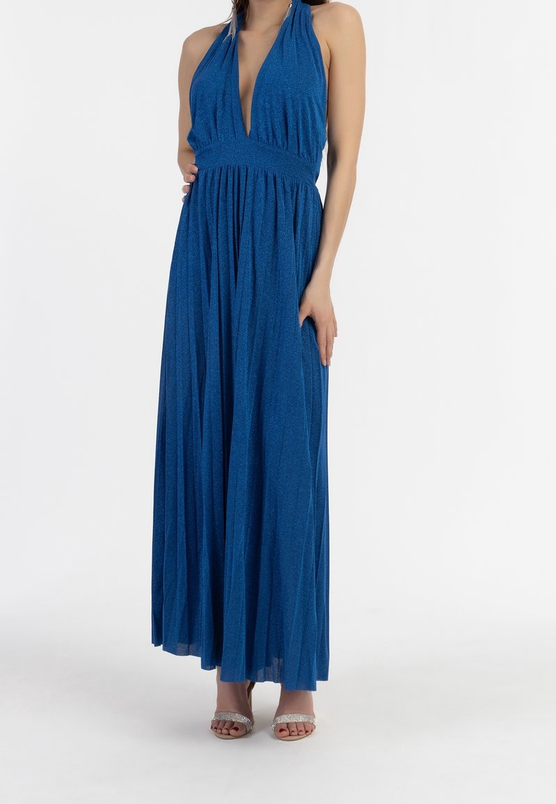 Robe maxi bleu à encolure halter, avec un décolleté plongeant, une jupe plissée et un tissu texturé. Associée à des sandales à talons argentées.