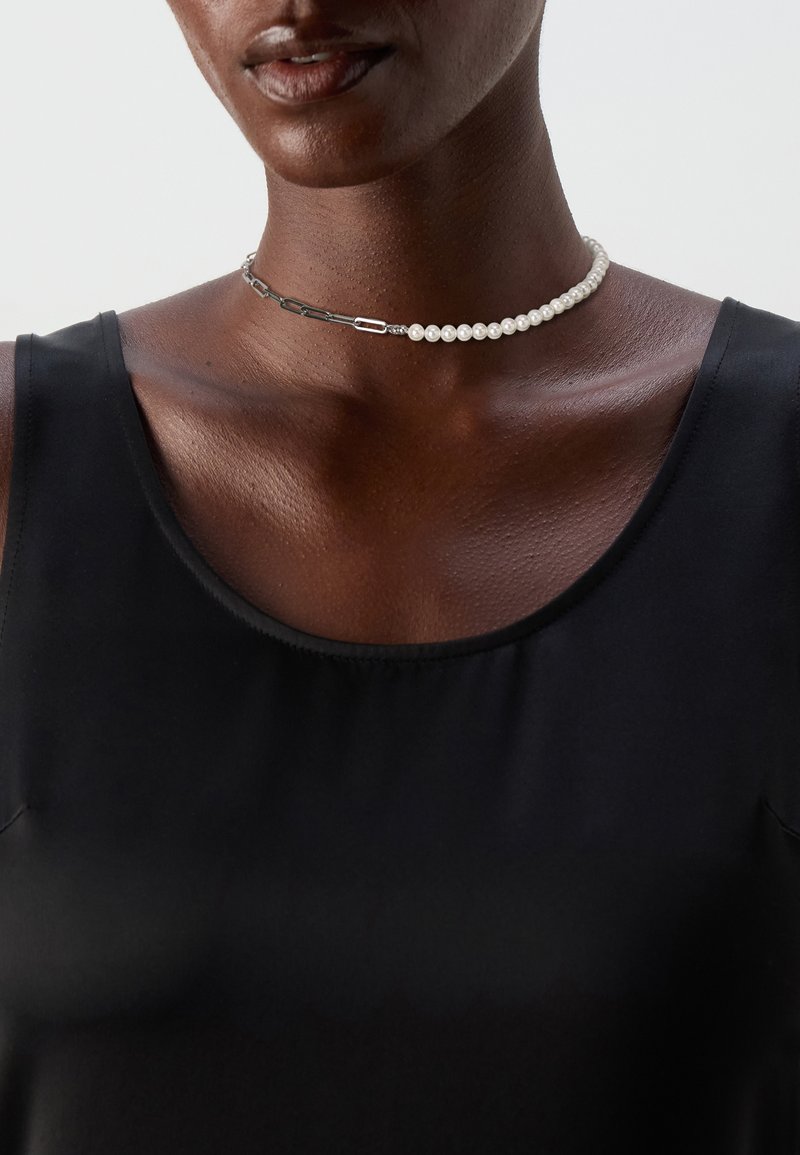 Lähikuva henkilöstä, jolla on musta hihaton toppi ja puoliksi helmiä, puoliksi metalliketjua oleva choker-kaulakoru.