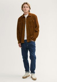 Brown corduroy jacket with two chest pockets, worn over a white t-shirt. Paired with blue denim jeans and cream sneakers with tan accents.