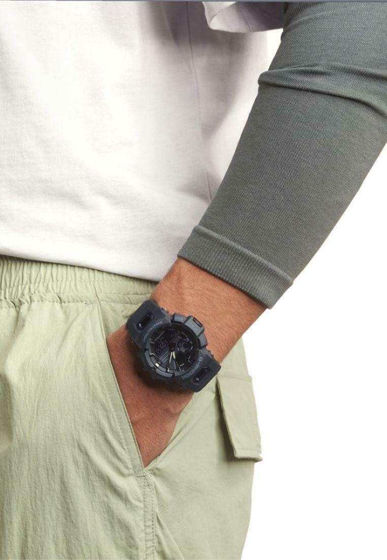Montre G-Shock noire au design robuste, avec un bracelet texturé et un grand cadran. Portée au poignet, associée à un vêtement vert clair et une manche grise.