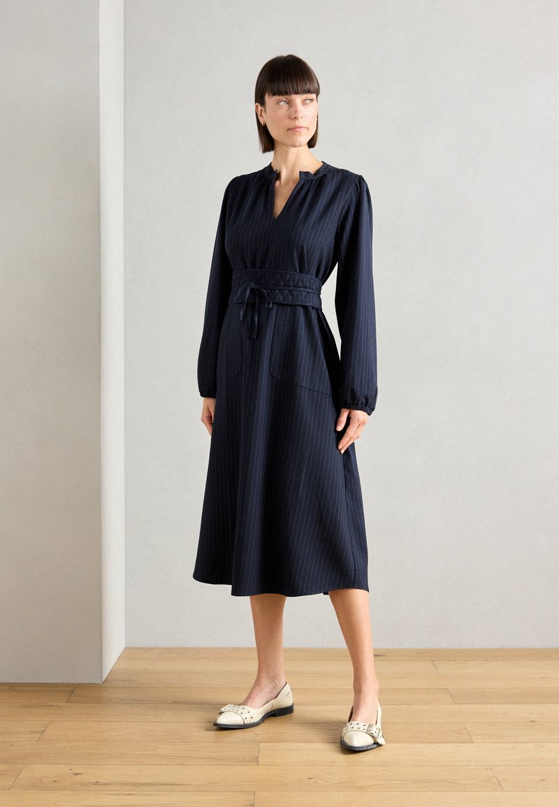Robe midi marine avec des rayures verticales, une taille plissée et des manches longues. Caractéristiques : poches latérales et accompagnée de ballerines crème.