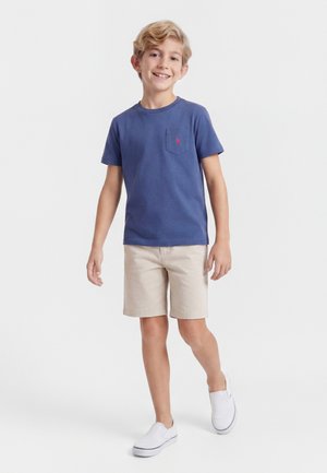 Χαμογελαστό αγόρι που περπατάει, φοράει μπλε κοντομάνικο πουκάμισο, μπεζ σορτς και λευκά παντόφλες, με φόντο απλό λευκό.