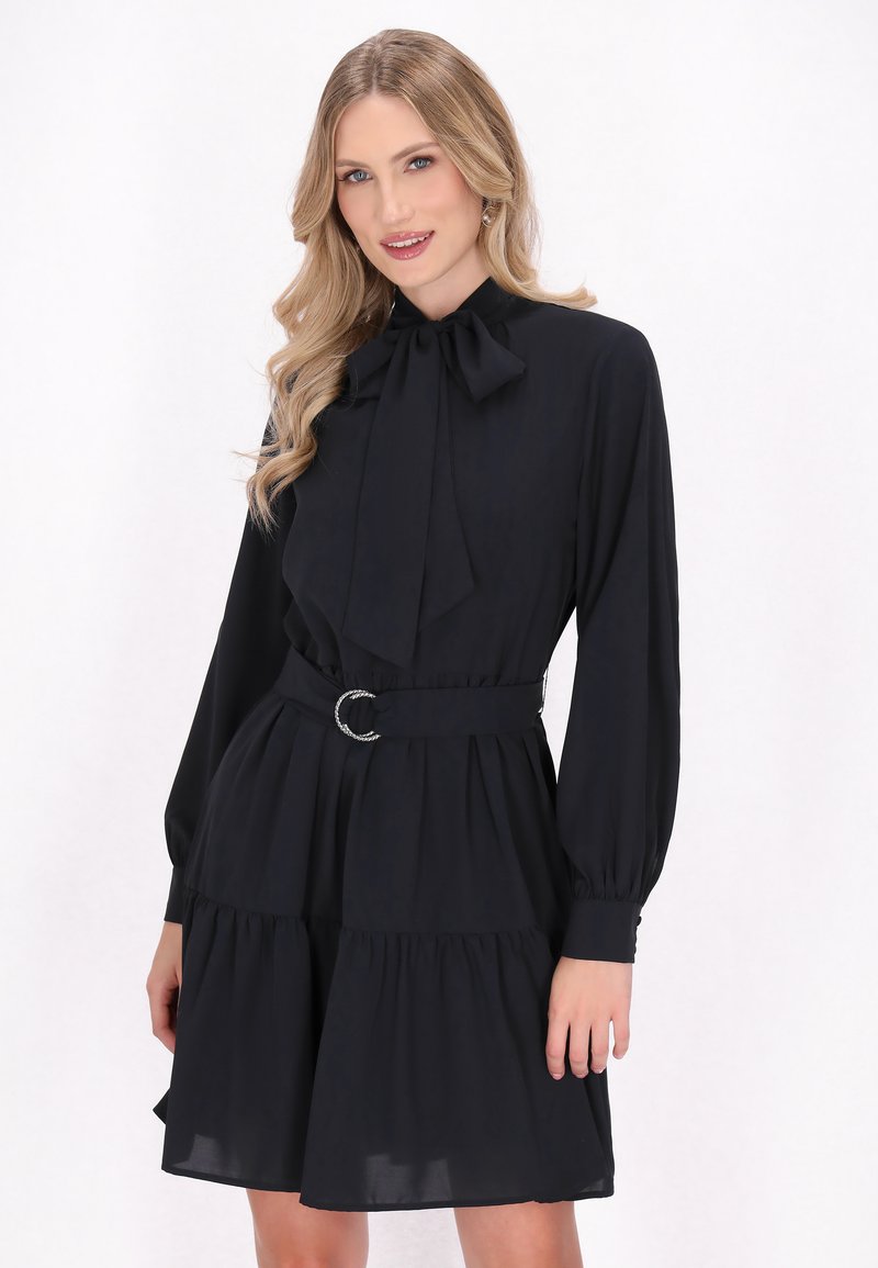 Robe noire à manches longues avec un col montant et un détail en nœud, cintrée à la taille avec une boucle en argent circulaire, présentant une jupe plissée.