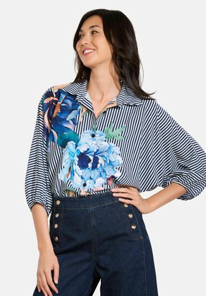 Camicia button-up con motivi floreali blu su uno sfondo a righe, colletto e maniche svasate. Abbinata a jeans scuri a vita alta.