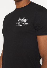 Czarna koszulka z krótkim rękawem z białym napisem "Replay Blue Jeans Authentic", noszona przez osobę o średnio-ciemnej karnacji.