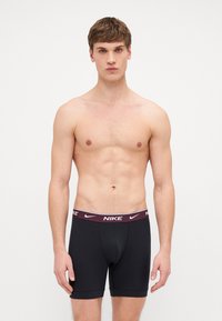 Juodos Nike šortai su burgundišku elastiniu diržu ir baltu logotipu. Audinys atrodo lygus ir prigludęs, pabrėžiantis klasikinį dizainą.