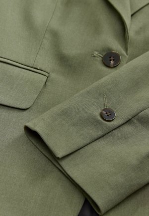 Olive green blazer with a textured fabric, featuring a single pocket and two brown buttons on the cuff, highlighting tailored design elements.