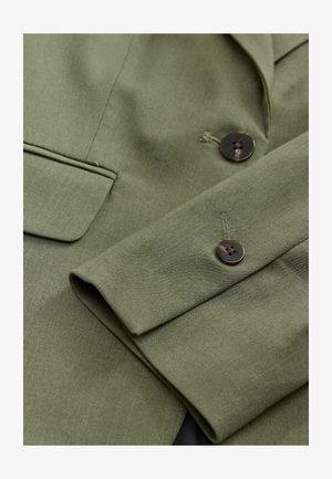 Olijfgroene blazer van een gestructureerde stof, met een enkele zak en twee bruine knopen op de mouw, die de op maat gemaakte designelementen benadrukt.