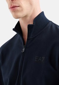 Giacca zip-up navy in morbido tessuto. Presenta un colletto alto, un logo discreto sul petto e una texture liscia.