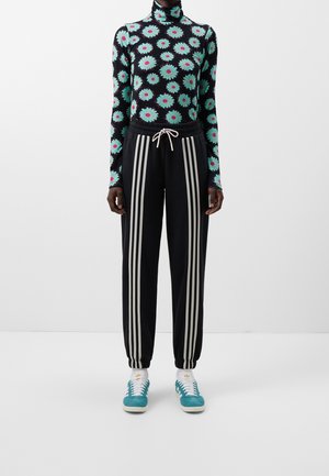 Henkilö pukeutuneena mustaan pitkähihainen puseroon, jossa on suuria turkooseja kukkia, mustat verkkarit valkoisilla raidoilla, valkoiset sukat ja turkoosit lenkkarit.