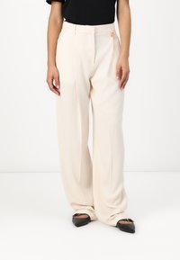 Beige wide-leg trousers with a smooth texture, featuring a single pleat and a gold-tone accent at the waist. Worn with black shoes.