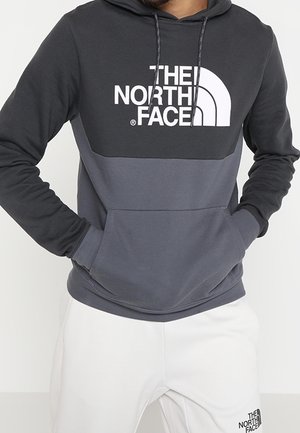 Osoba nosząca dwukolorową czarno-szarą bluzę z kapturem The North Face z rękami w przedniej kieszeni oraz białe spodnie z logo na lewym udzie.