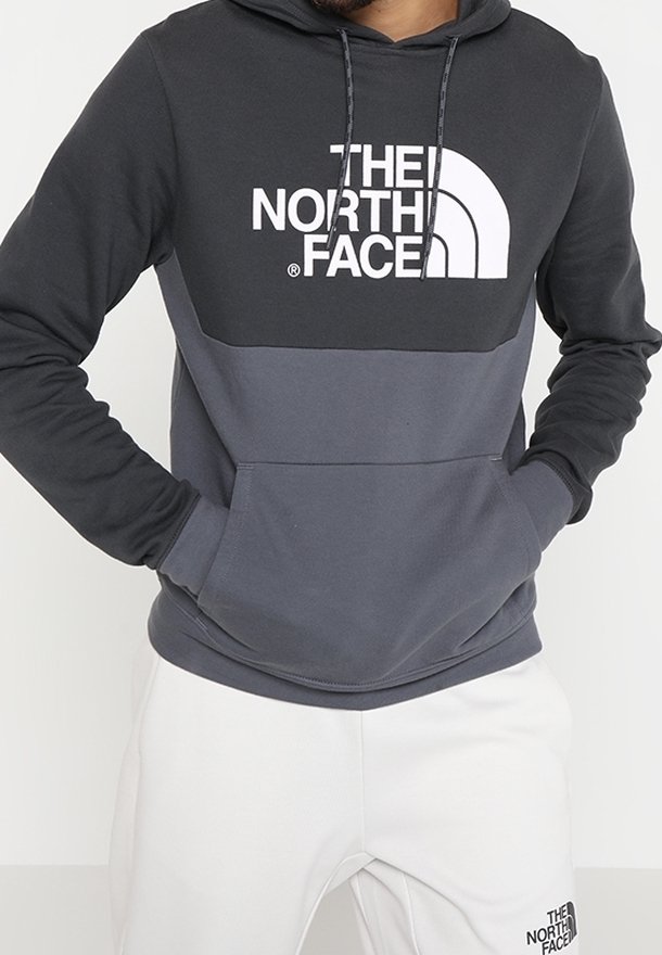 Personne portant un sweat à capuche The North Face bicolore noir et gris avec les mains dans la poche avant et un pantalon blanc avec un logo sur la cuisse gauche.