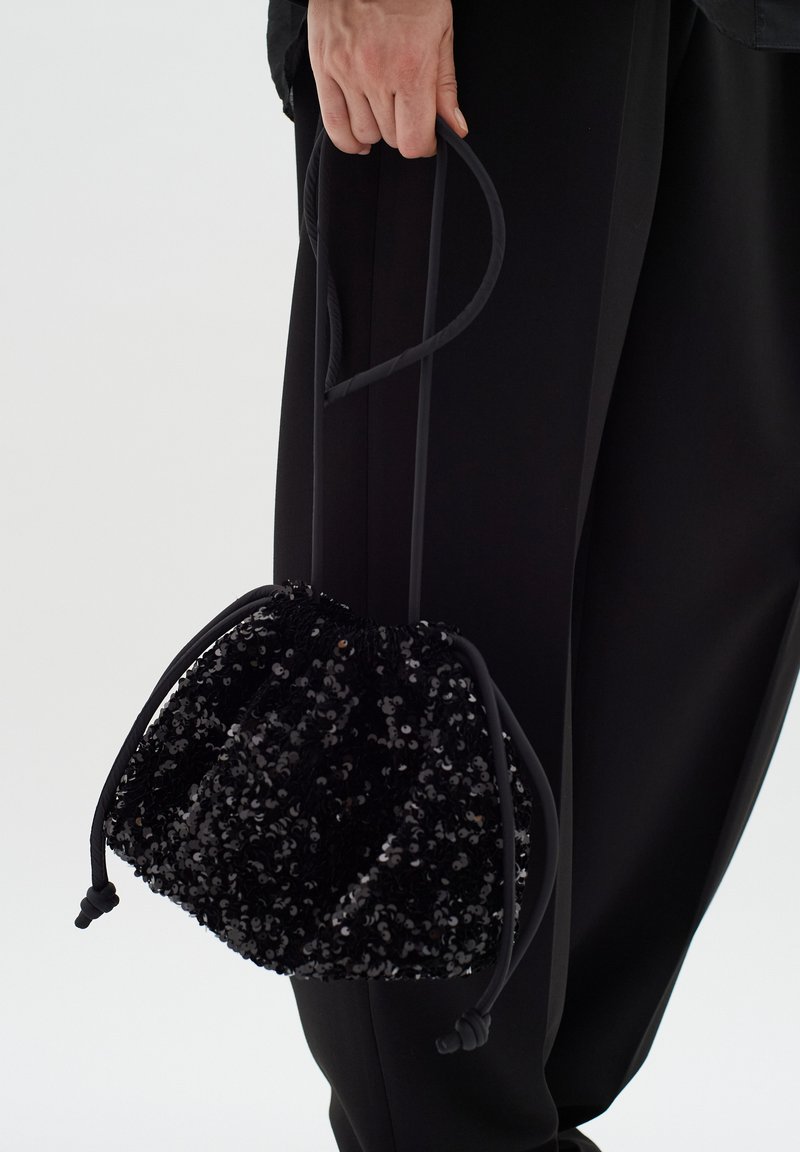 Bolsa de drawstring negra con lentejuelas y superficie texturizada, sostenida por una mano. Cintas ajustables negras y parte superior fruncida para el cierre.
