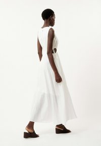 Robe maxi blanche fluide avec un décolleté en scoop et des découpes sur les côtés, maintenue par des cordons de serrage. Associée à des sandales à talons en cuir tressé marron.