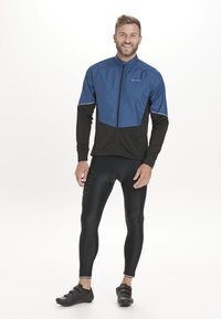 Herren-Radjacke in Blau und Schwarz, hergestellt aus leichtem, strukturiertem Material. Figurbetontes Design mit einem Reißverschluss und reflektierenden Akzenten. Schwarze Radhosen.