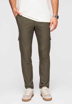 Mand iført olivengrønne cargobukser med klaplommer og beige sneakers, stående med den ene hånd i lommen og med synligt ur på håndleddet.