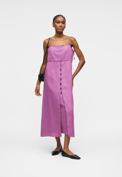 Femme portant une robe midi lavande sans manches avec un décolleté festonné, des découpes à l'avant, des ballerines noires et un petit sac à bandoulière noir.