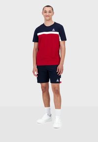 Röd och marinblå kortärmad t-shirt med vit rand, ihop med marinblå shorts. Har logotyp på bröstet och shorts, kompletterat med vita sneakers.