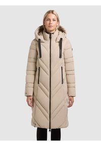 Lange beige pufferjas met een capuchon, voorzien van een chevron-gestikt patroon, een zwarte rits en verstelbare koordjes op de capuchon.