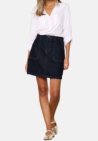 Kvinna som bär en vit och rosa randig blus instopad i en mörk denimkjol med framfickor och metallicguld Mary Jane-skor.