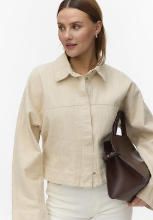 Frau trägt eine beige strukturierte Jacke und weiße Hose, hält eine dunkelbraune trapezförmige Handtasche und schaut nach links.