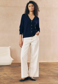 Marinblå cardigan med knappar, V-ringning och långa ärmar som matchas med vita vida byxor. Svarta platta skor fulländar outfiten.