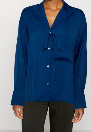 Blusa di raso blu scuro con un nodo al colletto, maniche lunghe e una tasca frontale. Presenta bottoni lungo la patta e una texture liscia.