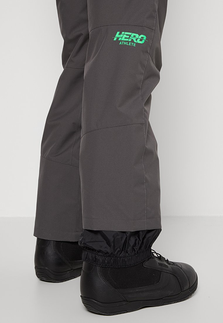 Szare spodnie sportowe o taktycznym wzornictwie, z elastycznymi mankietami i zielonym logo. Noszone z czarnymi butami outdoorowymi, podkreślającymi ich funkcjonalność.