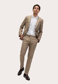 Beiger, maßgeschneiderter Anzug mit tailliertem Sakko und schmalen Hosen, kombiniert mit einem weißen Hemd. Braune Schuhe komplettieren den Look. Glatte Stoffstruktur.