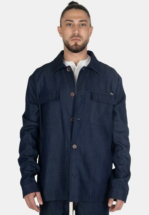 Uomo con la barba che indossa una giacca di jeans blu scuro abbottonata e una camicia bianca davanti a uno sfondo chiaro e semplice.