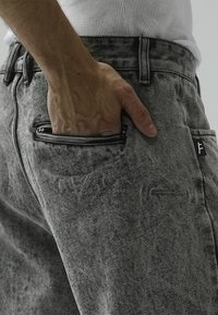 Grå denimjeans har en slank passform med en teksturert finish. En hånd er plassert i en frontlomme, noe som viser et stripete innerfôr.