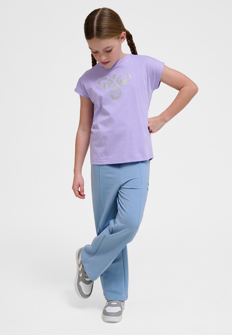 Lila T-Shirt mit kurzen Ärmeln und silbernem Grafikdesign, kombiniert mit hellblauen Jogginghosen. Die Schuhe sind grau mit weißen Akzenten.