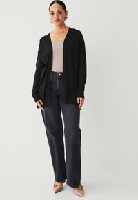 Zwarte cardigan met open voorzijde, gedragen over een lichte tanktop, gecombineerd met donkere jeans en nude puntige hakken. Gladde textuur.