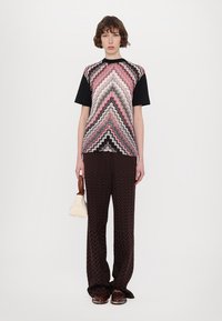 Camiseta de patrón zigzag multicolor con mangas cortas negras, combinada con pantalones de pierna ancha en patrón zigzag marrón y un pequeño bolso de crema.