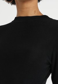 Gros plan sur une personne portant un haut à manches longues noir côtelé avec un col rond et une petite tache de naissance sur le cou.