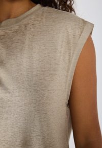 Beige ermeløs topp laget av teksturert stoff, med rund halslinje og lett rullede skuldersømmer. Glatt og lett utseende.